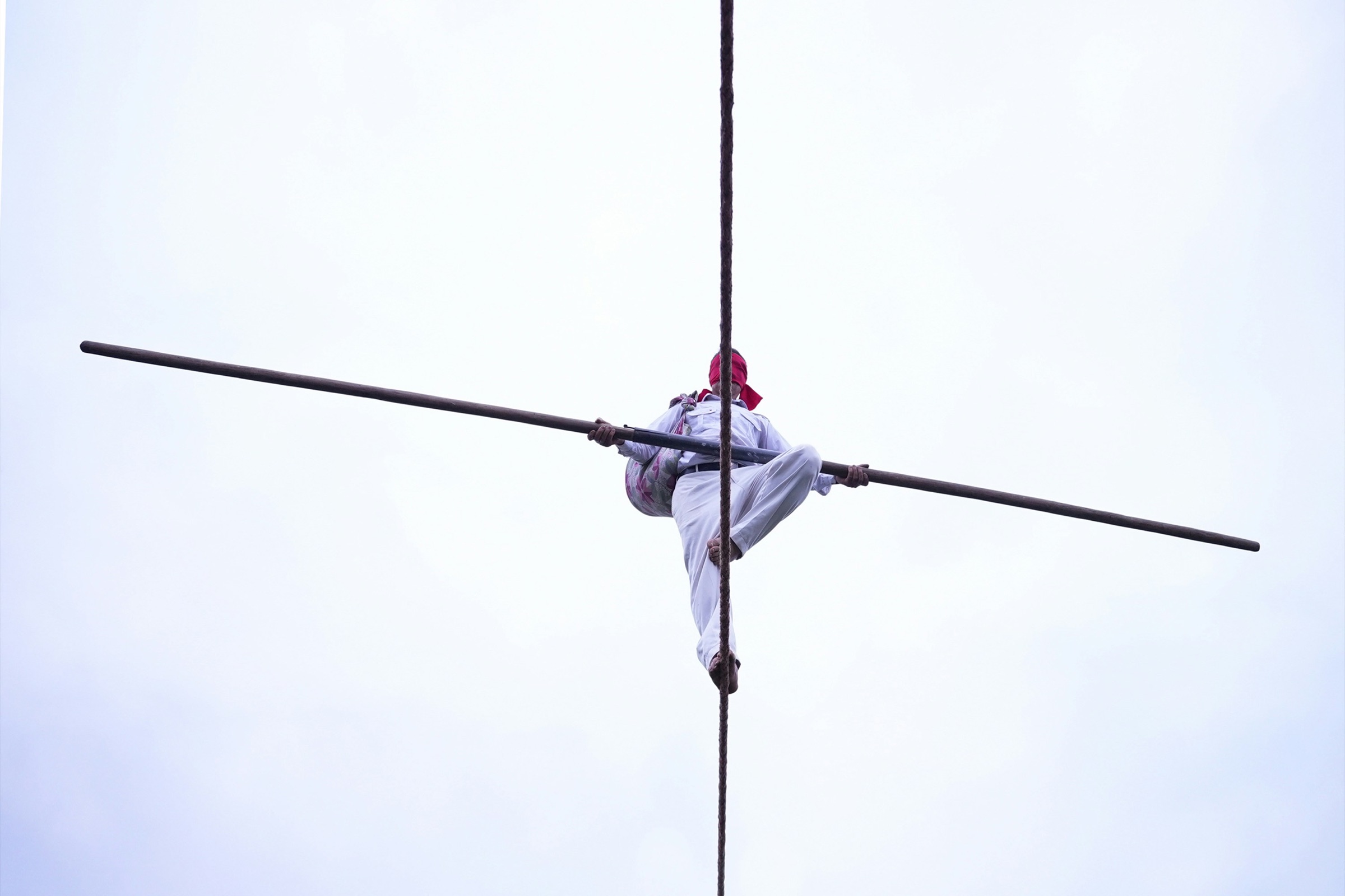A rope walker high against an empty pale sky, seen from below. The rope runs vertically and the balance pole runs horizontally, crossing at the figure.